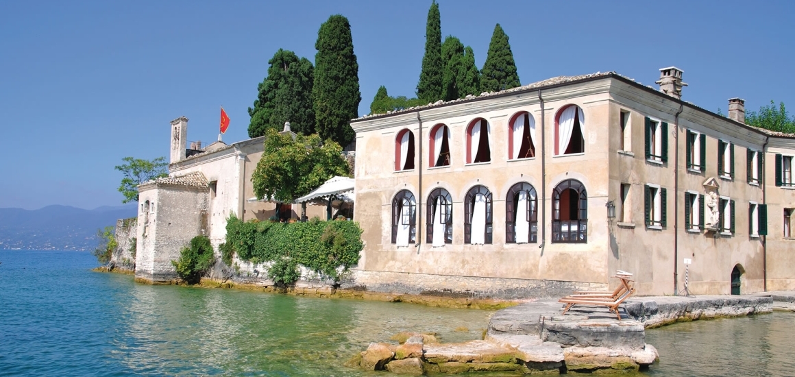 Historisches Gebäude am Ufer des Gardasees in San Vigilio, umgeben von Wasser und Bäumen.