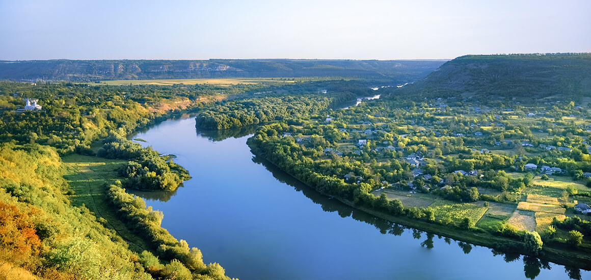 Landschaft mit einem Fluss, der sich durch eine grüne, bewaldete Gegend schlängelt, umgeben von Hügeln und kleinen Siedlungen.