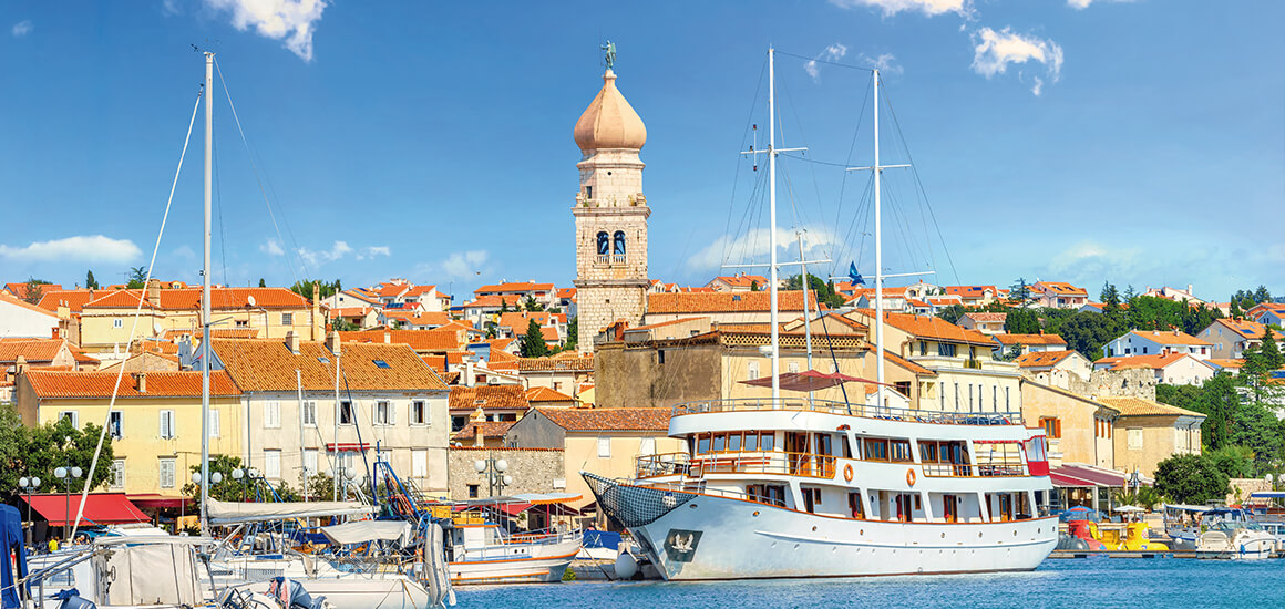 Panorama der Stadt Krk auf der Insel Krk in Kroatien mit einem großen Boot im Vordergrund und einer Kirche mit einem markanten Turm im Hintergrund.