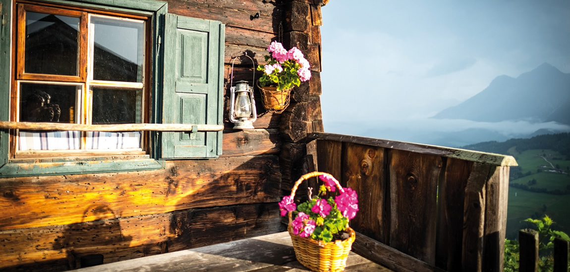 Ein rustikales Holzhaus mit einem Fenster und grünen Fensterläden, daneben eine Laterne und ein Blumenkorb mit rosa Blumen. Im Hintergrund sind Berge und eine neblige Landschaft zu sehen.