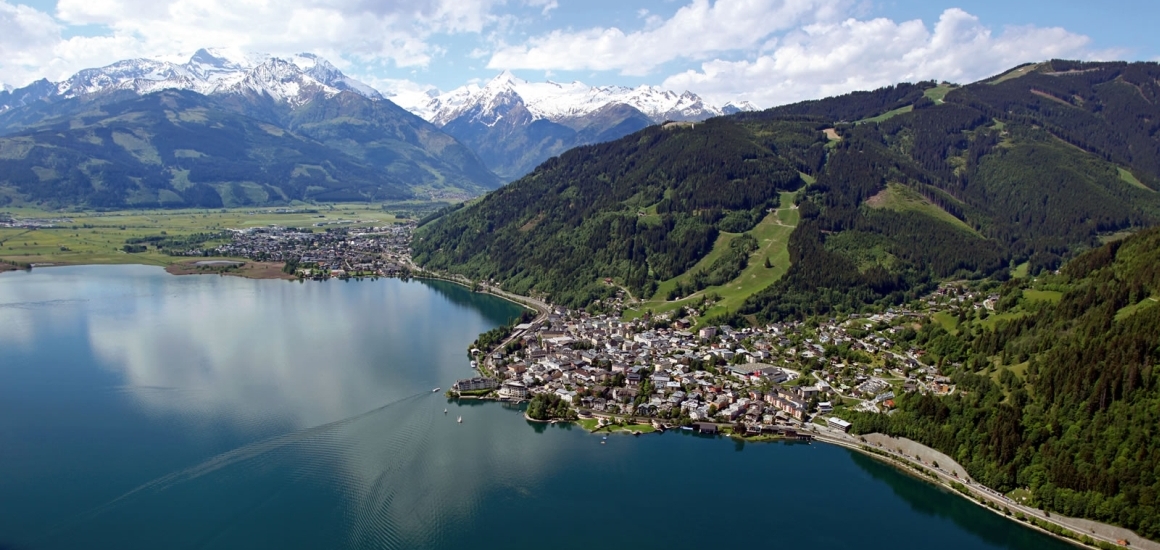 Luftaufnahme von Zell am See mit dem Zeller See im Vordergrund und den Alpen im Hintergrund.