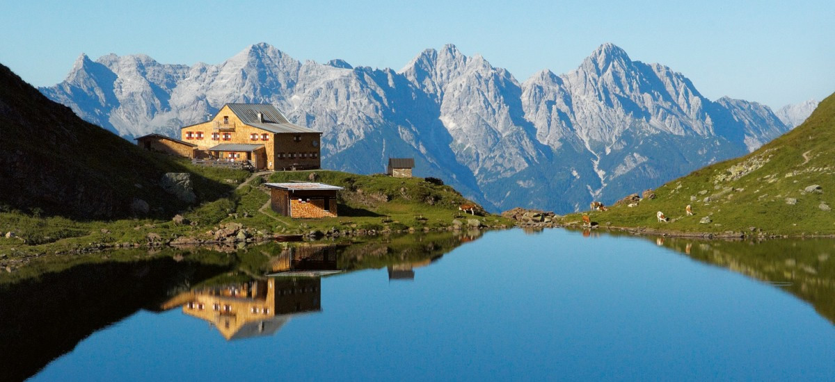 Ein Bergsee in Tirol mit einem Gebäude am Ufer und Bergen im Hintergrund, die sich im Wasser spiegeln.