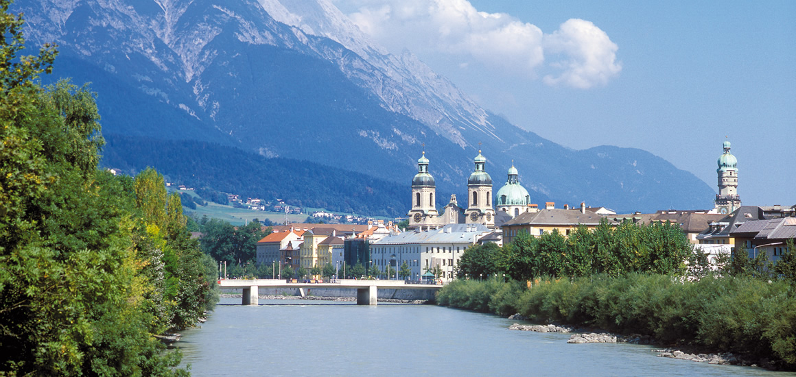 Blick auf Innsbruck mit dem Karwendelgebirge im Hintergrund, Tirol, Österreich. Im Vordergrund fließt ein Fluss, umgeben von Bäumen und Gebäuden mit markanten Kirchtürmen.