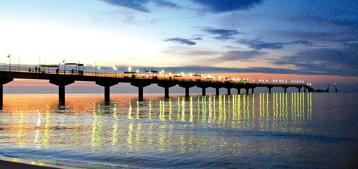 Außenansicht der Misdroy Seebrücke mit Menschen im Vordergrund, begrünten Bereichen und modernen Strukturen.