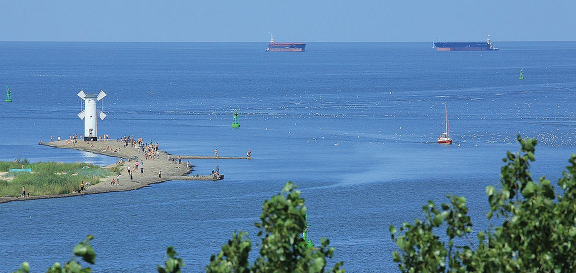 Blick auf den Leuchtturm in Swinemünde am Meer mit mehreren Schiffen im Hintergrund.