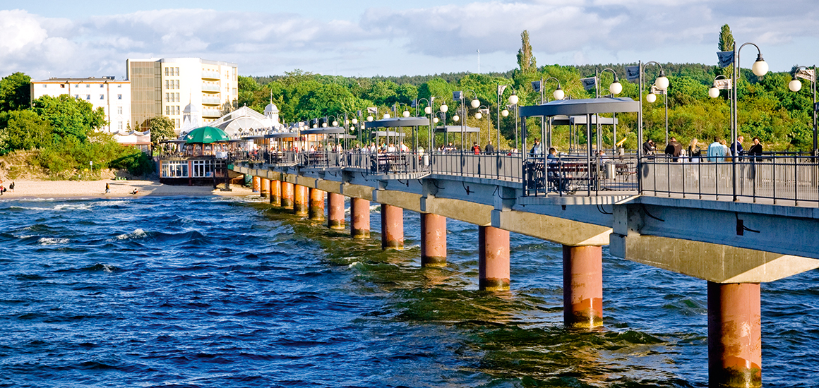 Seebrücke in Swinemünde, Polen, mit Menschen, die darauf spazieren gehen. Die Brücke erstreckt sich über das Meer, umgeben von grüner Landschaft und Gebäuden im Hintergrund.