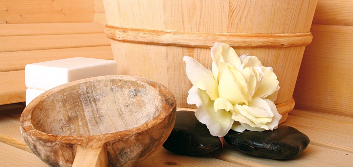 Ein Sauna-Set mit einem hölzernen Aufgusskübel, einer Kelle, Seifenstücken und einer Blume, arrangiert auf einer Holzoberfläche.