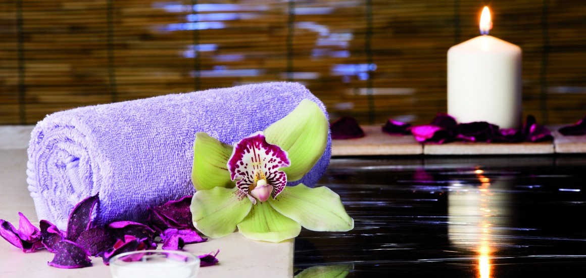 Ein Wellness-Stilleben mit einem lila Handtuch, einer grünen Blume, lila Blütenblättern, einer brennenden Kerze und einem Wasserbecken.