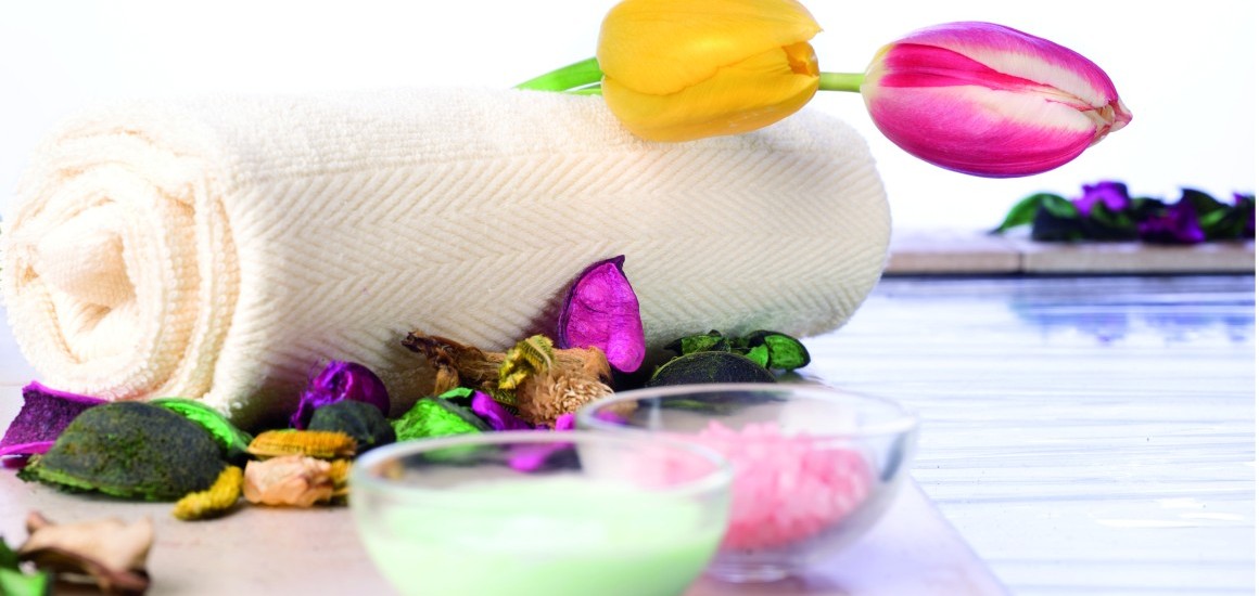Ein Stillleben mit einem zusammengerollten Handtuch, einer gelben und einer rosa Tulpe, umgeben von bunten Blütenblättern und zwei kleinen Schalen mit grüner und rosa Substanz, auf einem Tisch arrangiert.