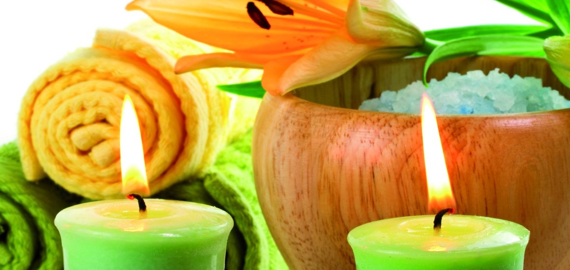 Ein Stillleben mit zwei brennenden grünen Kerzen, einer orangefarbenen Lilie, einem Holzgefäß mit Badesalz und einem gelben Handtuch im Hintergrund.