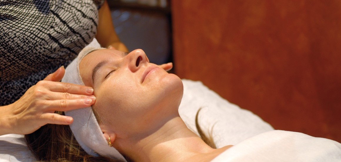 Eine Person liegt entspannt auf einer Wellness-Liege und erhält eine Gesichtsmassage. Die Umgebung wirkt beruhigend und entspannend.