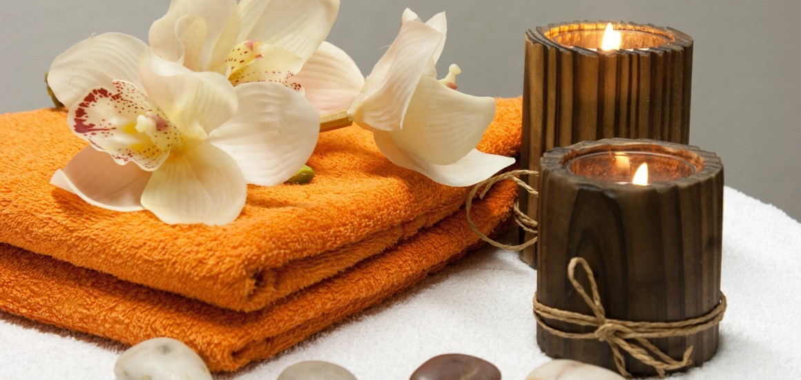 Ein Wellness-Arrangement mit zwei brennenden Kerzen, weißen Orchideenblüten, einem orangefarbenen Handtuch und glatten Steinen auf einem weißen Untergrund.