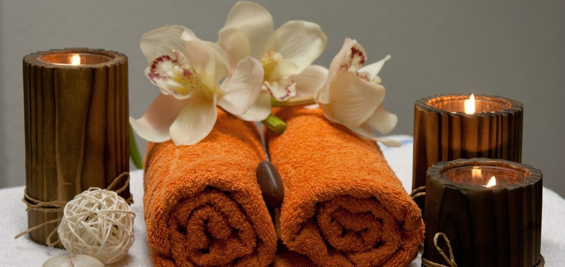Zwei orangefarbene Handtücher, dekoriert mit weißen Orchideenblüten, umgeben von brennenden Kerzen und einer dekorativen Kugel, auf einem Tisch arrangiert.