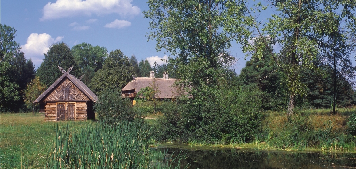 Landschaftsbild aus Masuren, Polen, mit traditionellen Holzhäusern, Bäumen und einem Teich im Vordergrund.
