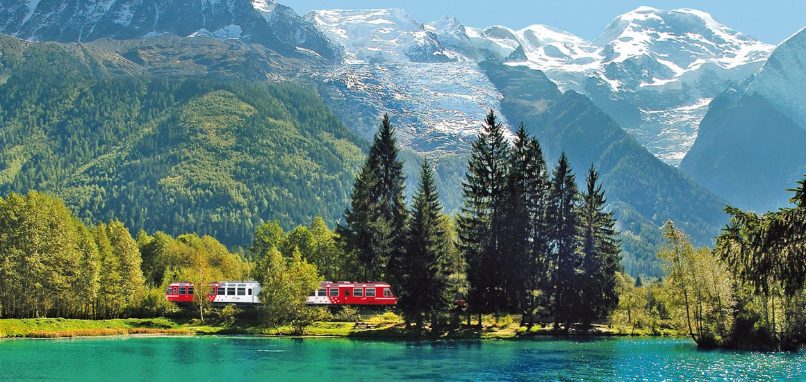 Ein roter Zug fährt durch eine malerische Landschaft mit einem See im Vordergrund, umgeben von Bäumen und Bergen im Hintergrund, die mit Schnee bedeckt sind.