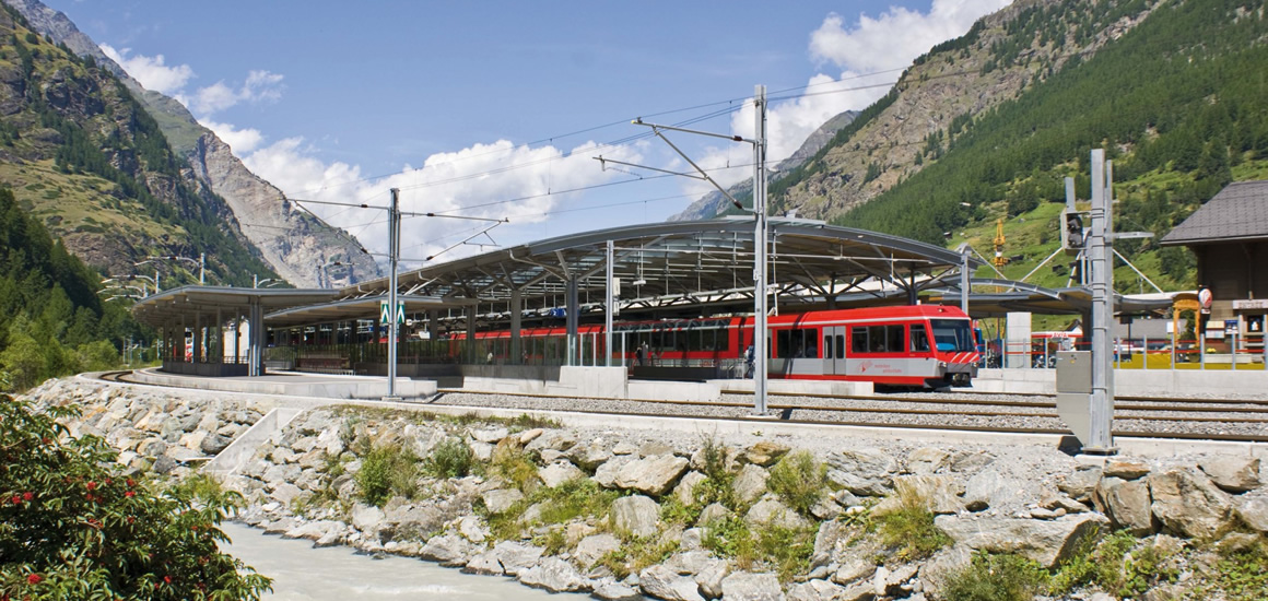 Bahnhof Täsch in der Schweiz mit einem roten Zug auf den Gleisen, umgeben von Bergen und einem Fluss im Vordergrund.