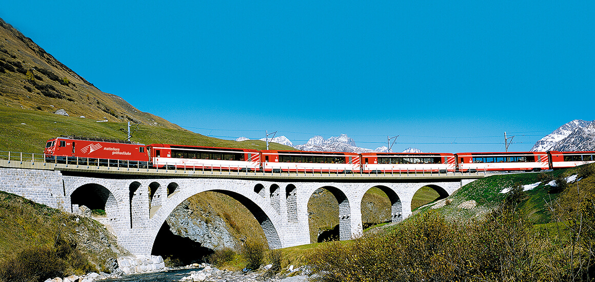 Ein roter Zug fährt über eine Steinbrücke in einer bergigen Landschaft mit klarem Himmel.