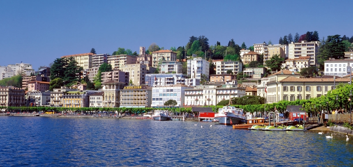 Panoramablick auf die Stadt Lugano in der Schweiz mit Gebäuden am Ufer des Luganersees und bewaldeten Hügeln im Hintergrund.