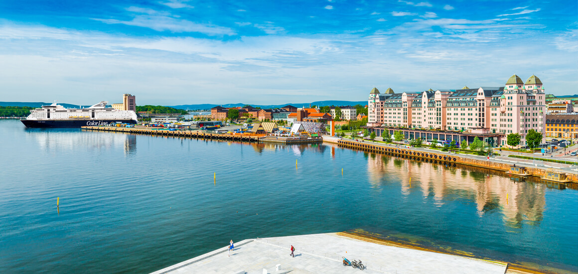 Panoramablick auf einen Hafen mit einem großen Kreuzfahrtschiff und einer Reihe von Gebäuden entlang der Küste, unter einem blauen Himmel.