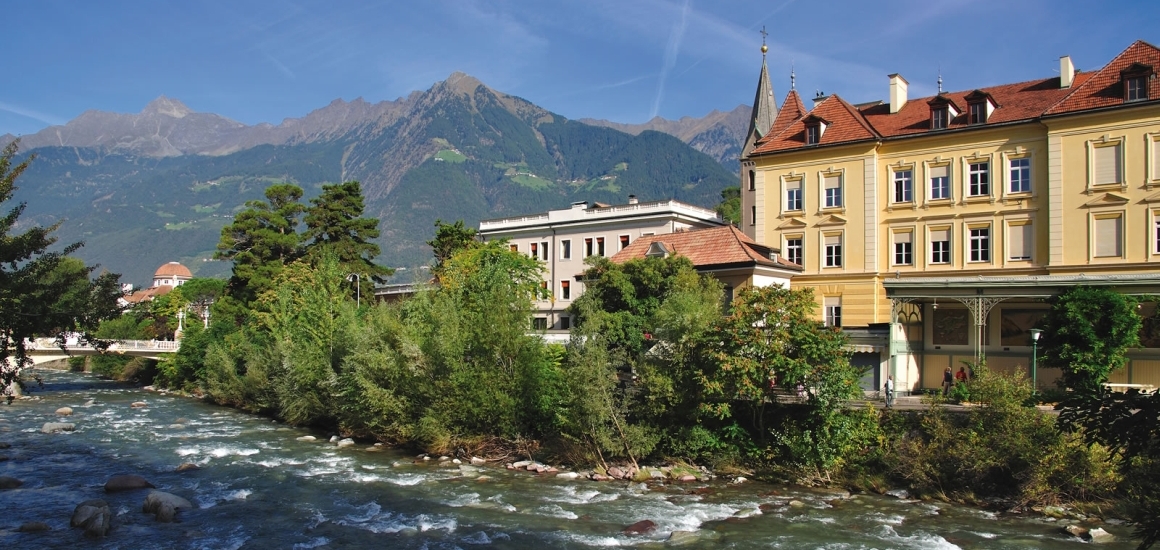 Blick auf Meran mit Fluss, Gebäuden und Bergen im Hintergrund.