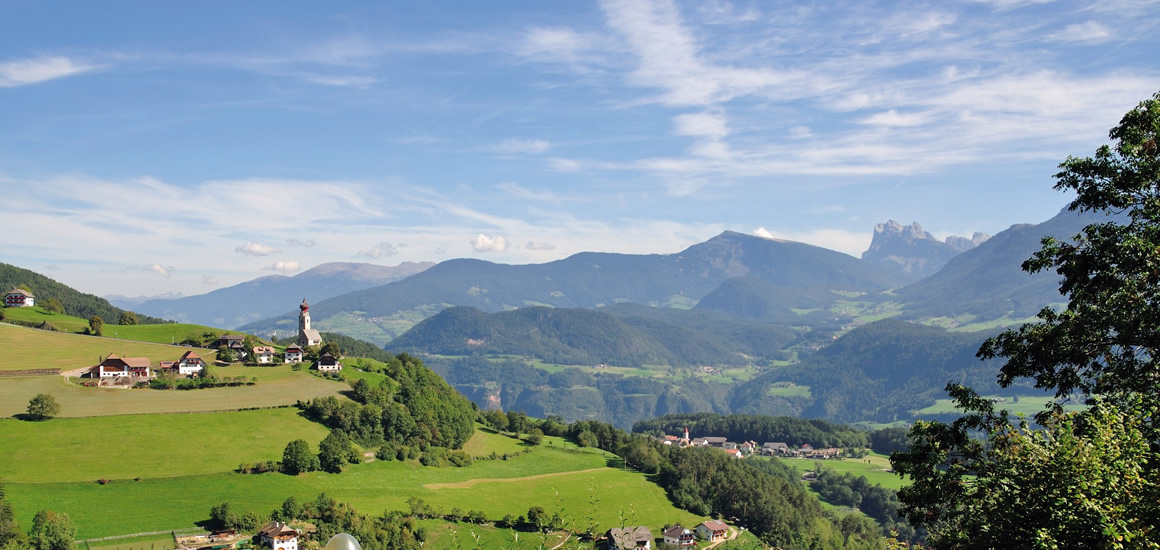 Landschaftsbild von Südtirol mit grünen Hügeln, einem Dorf mit Kirche und Bergpanorama im Hintergrund.
